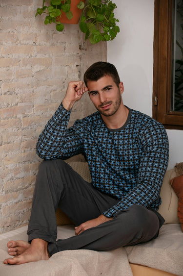 Man sitting on a couch wearing a blue patterned shirt and gray pants, with a plant and brick wall in the background.